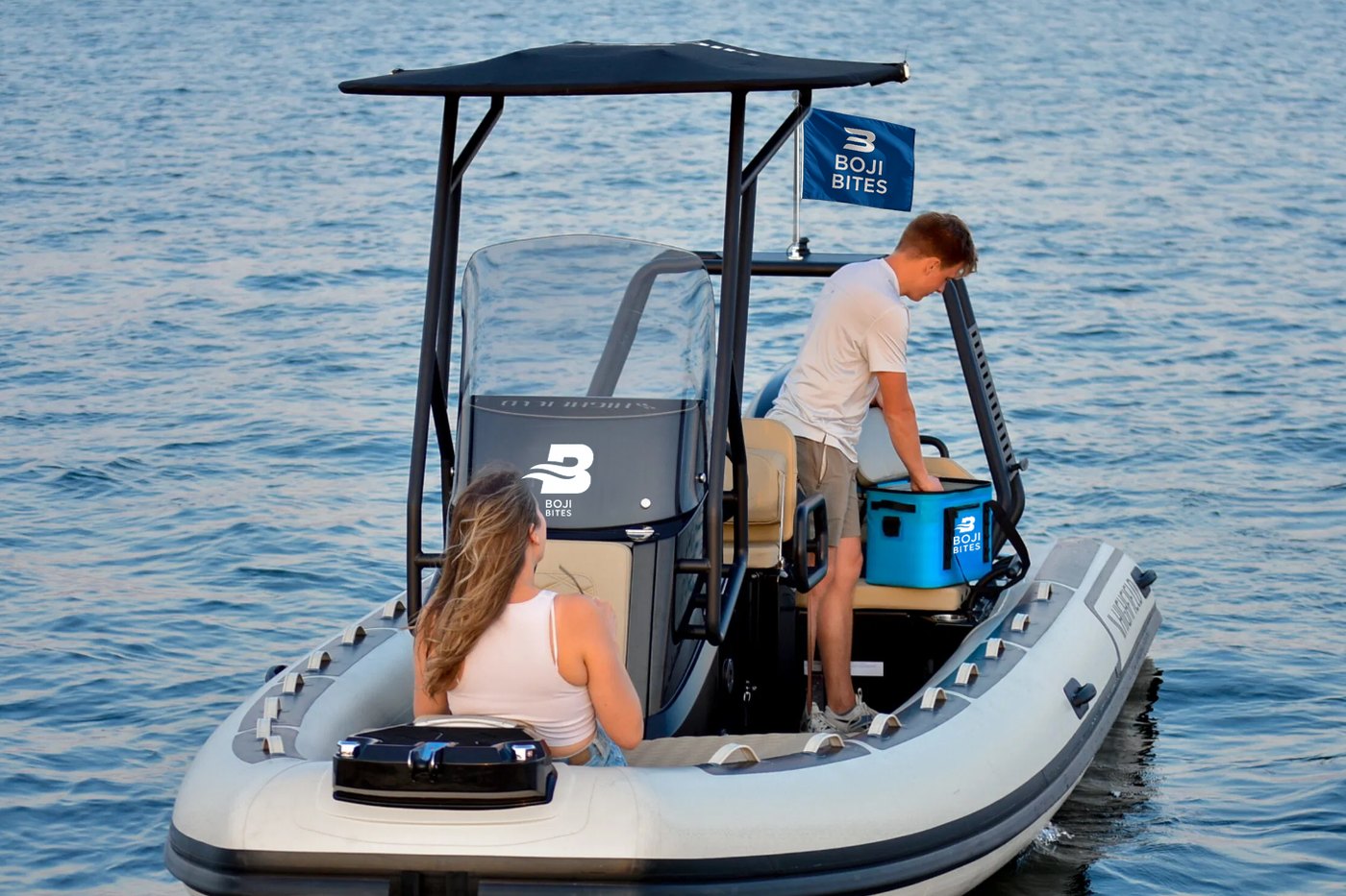 The Boji Bites Highfield RIB on West Okoboji at golden hour, blue Boji Bites flag flying from the T-top, crew loading a branded cooler aboard
